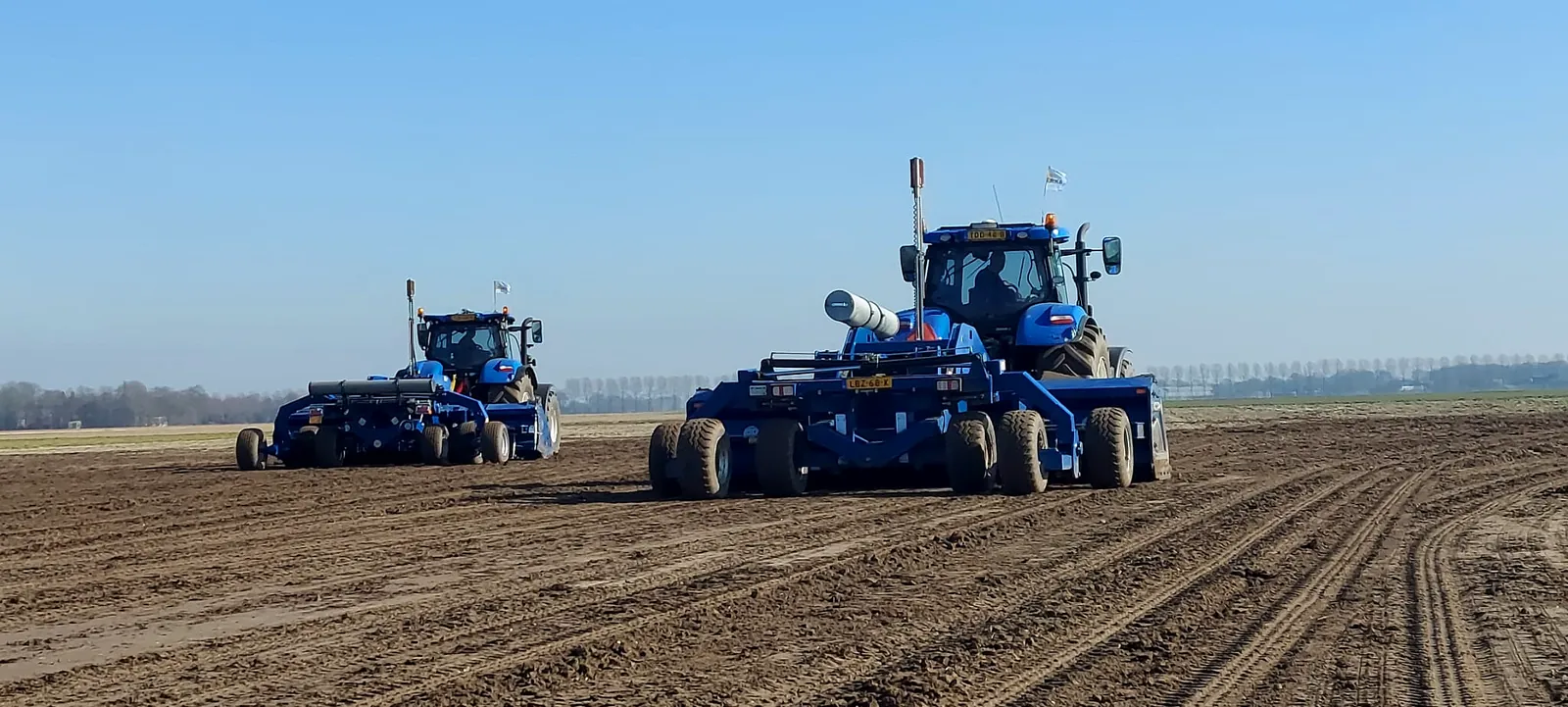 Twee New Holland trekkers met kilverbakken naast elkaar op een groot perceel