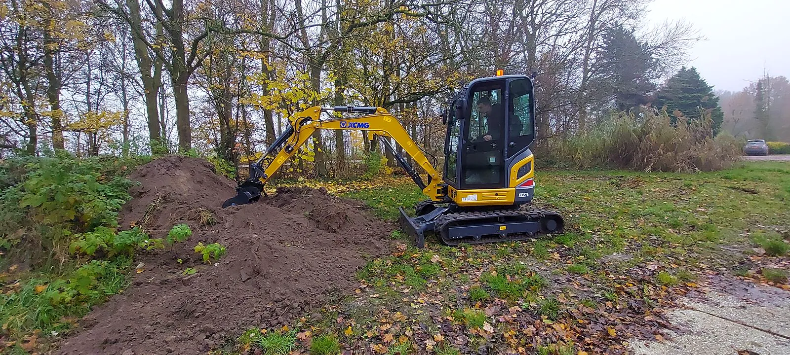 Minikraan bij een bomenwal met een hoop uitgegraven grond